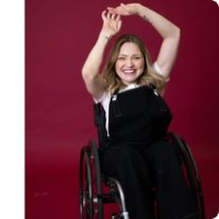 smiling woman in wheelchair raising her arms above her head wearing black dungarees